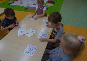 Dzieci siedzą przy stoliku. Chłopiec trzyma w ręku nożyczki i wycina małe znaki drogowe. Dwoje dzieci układa sudoku z wyciętych znaków, nakleja je we wlaściwe miejsce. Jedna dziewczynka przygląda się ułożonemu sudoku.